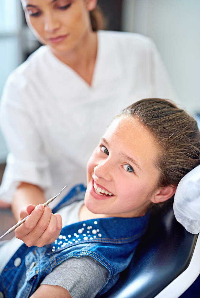 Young girl smiling in a dental chair while holding a dental tool, with a dental professional standing nearby – DeMoss Pediatric Dentistry | Dentist Englewood Ohio